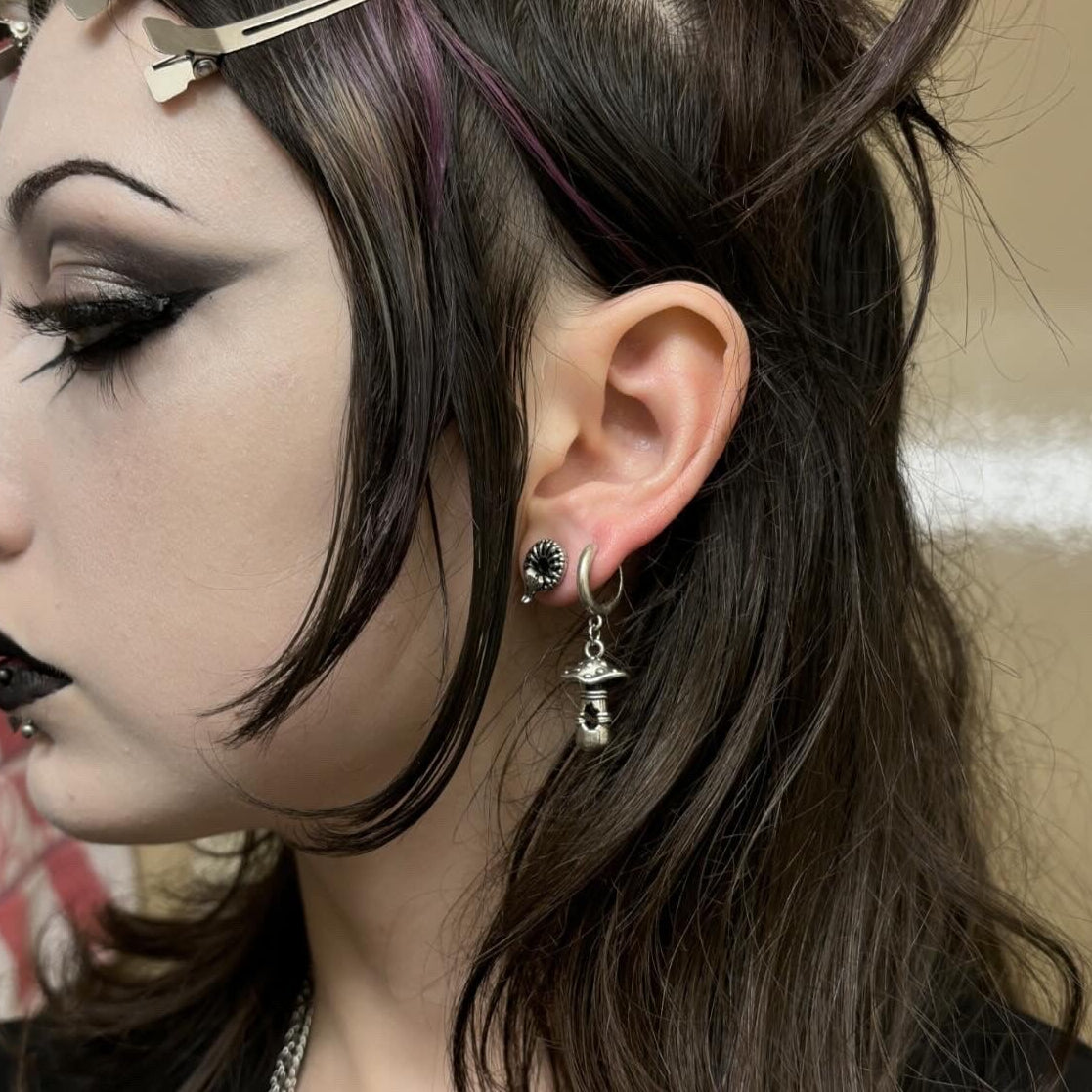 Side profile of a person with dark makeup wearing a hoop earring with a mushroom charm and a stud earring