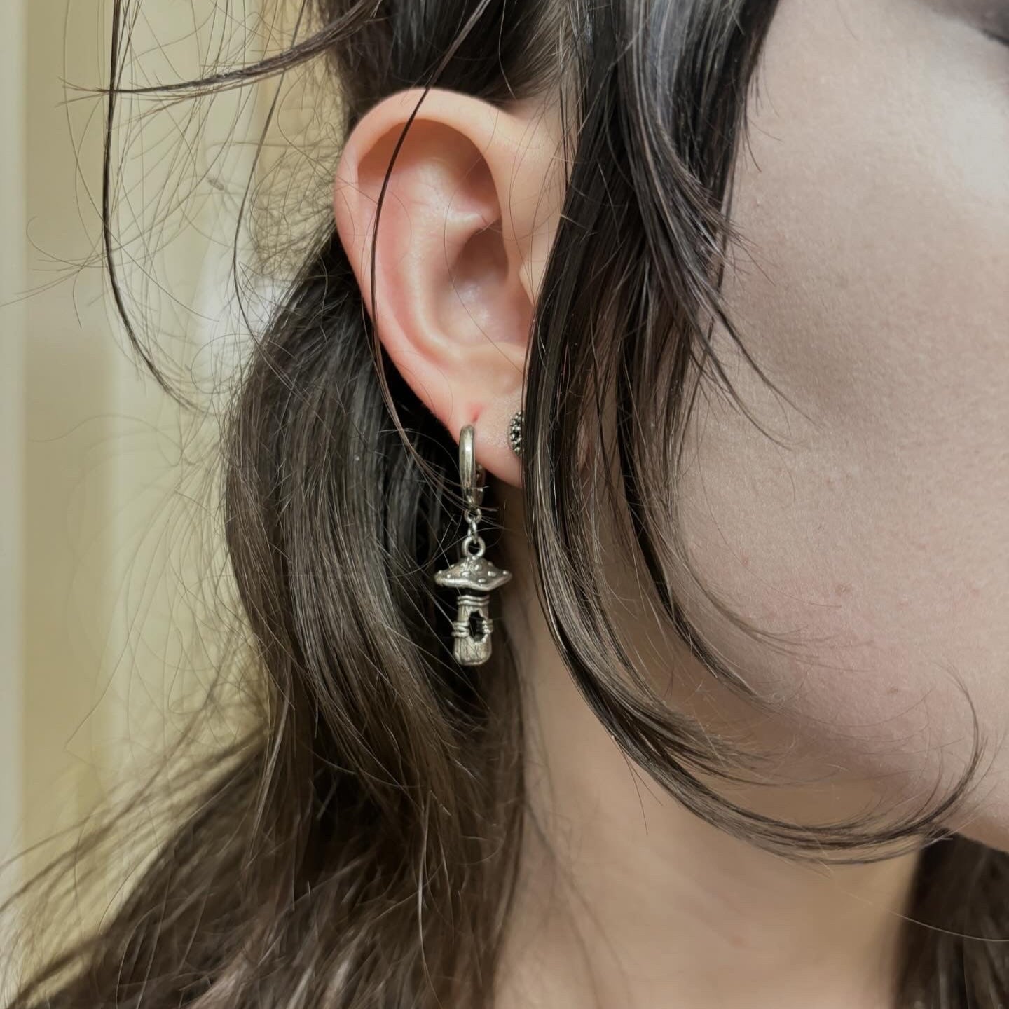 Close-up of a small hoop earring with a mushroom charm worn on a person's ear beneath loose brown hair