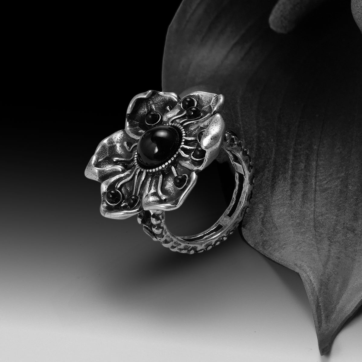 Silver-tone flower ring with black stone center resting against a dark leaf, close-up angled view