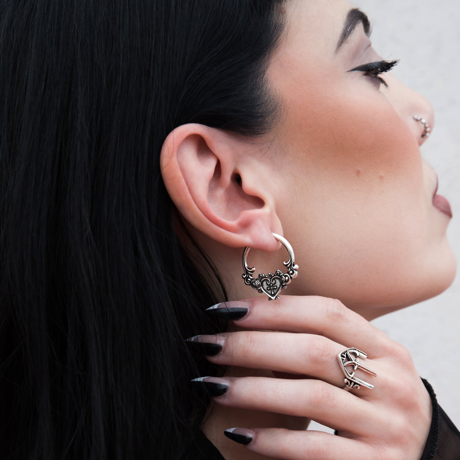 Silver-tone ornate hoop earring with heart and cross motif worn on a model's ear, close-up side view