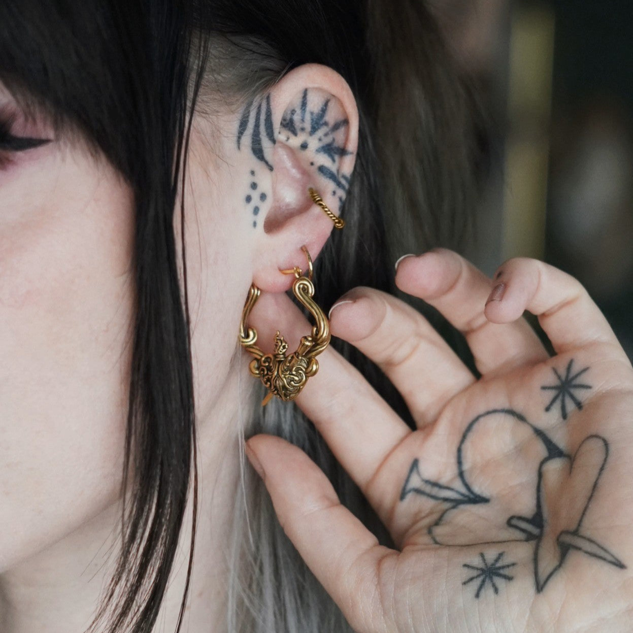 Person with tattooed hand touching a gold-tone flaming heart hoop earring worn in the ear