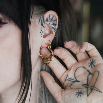 Person with tattooed hand touching a gold-tone flaming heart hoop earring worn in the ear