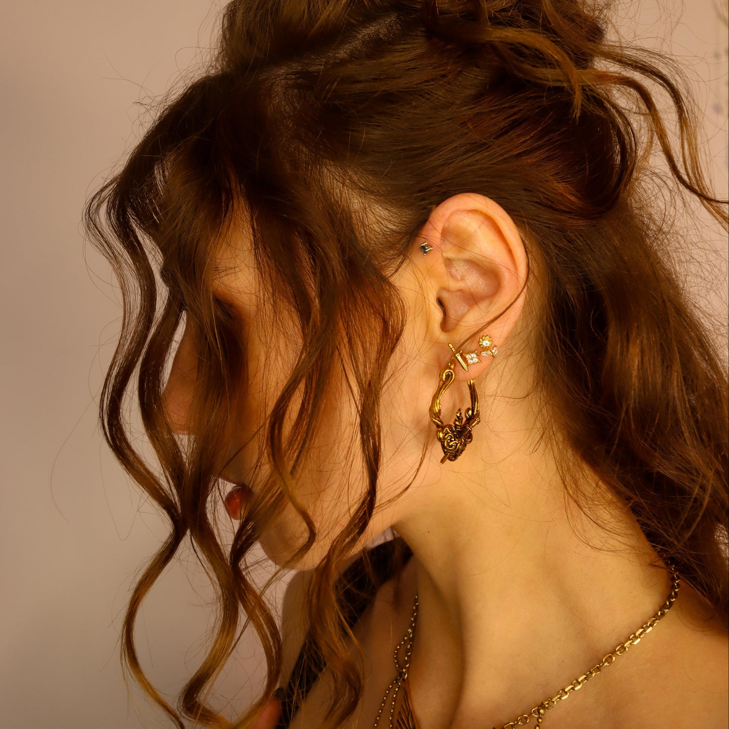 Side profile of a person with wavy hair wearing a gold-tone heart-shaped hoop earring among multiple ear piercings