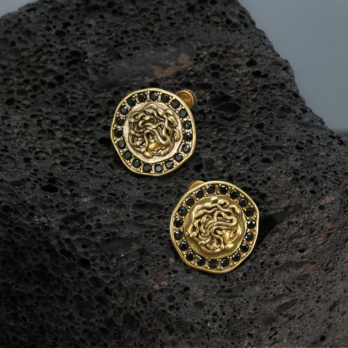 petrified nocturne ear studs in gold shown in a product view on black volcanic rock, featuring medusa-inspired ear studs framed with black cubic zirconia