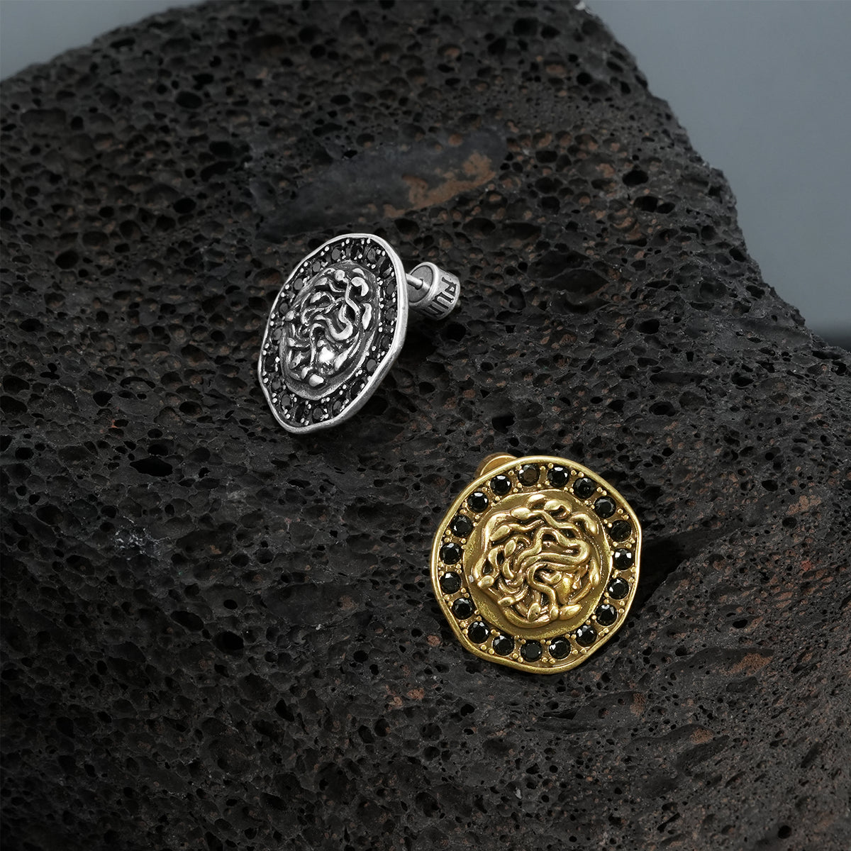 petrified nocturne ear studs shown in silver and gold in a product view on black volcanic rock, featuring medusa-inspired ear studs framed with black cubic zirconia