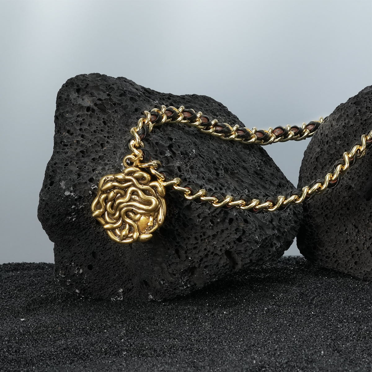 petrified power necklace in gold shown in a product view on black volcanic rock, featuring a medusa-inspired pendant on a braided chain