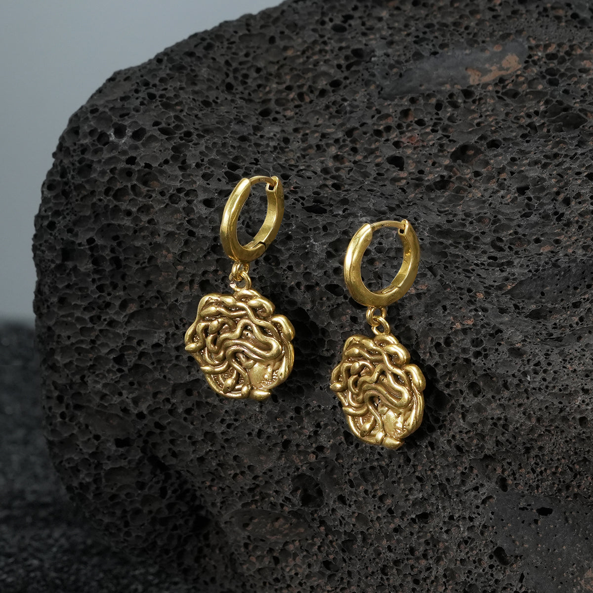 petrifying gaze dangle earrings in gold shown in a product view on black volcanic rock, featuring medusa-inspired charms suspended from hoops
