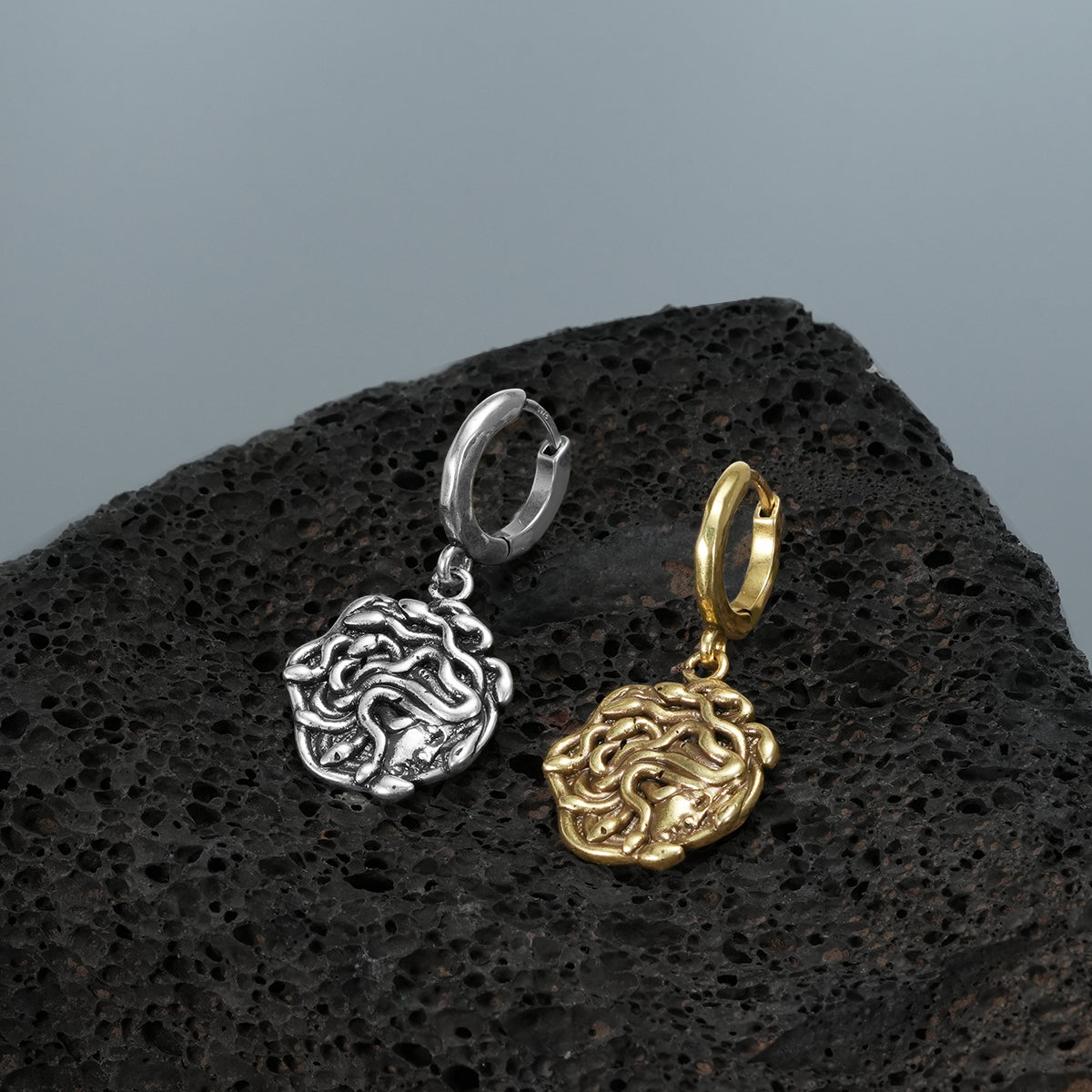 petrifying gaze dangle earrings shown in silver and gold in a product view on black volcanic rock, featuring medusa-inspired charms suspended from hoops