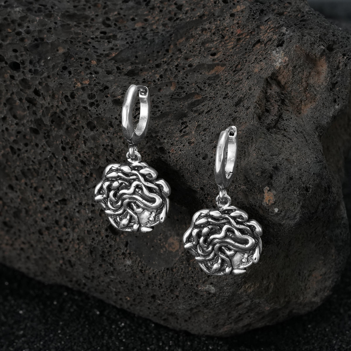 petrifying gaze dangle earrings in silver shown in a product view on black volcanic rock, featuring medusa-inspired charms suspended from hoops