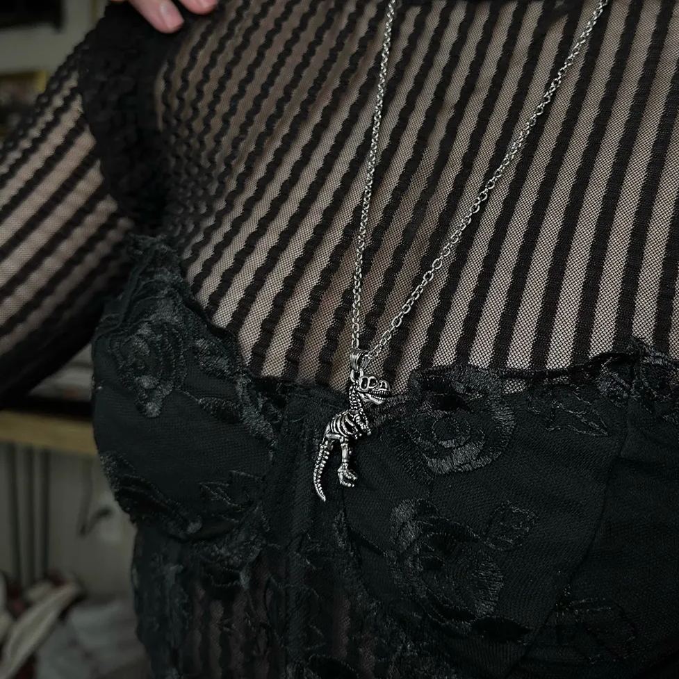 Close-up view of a silver-tone T-rex skeleton pendant necklace against a black sheer and lace top