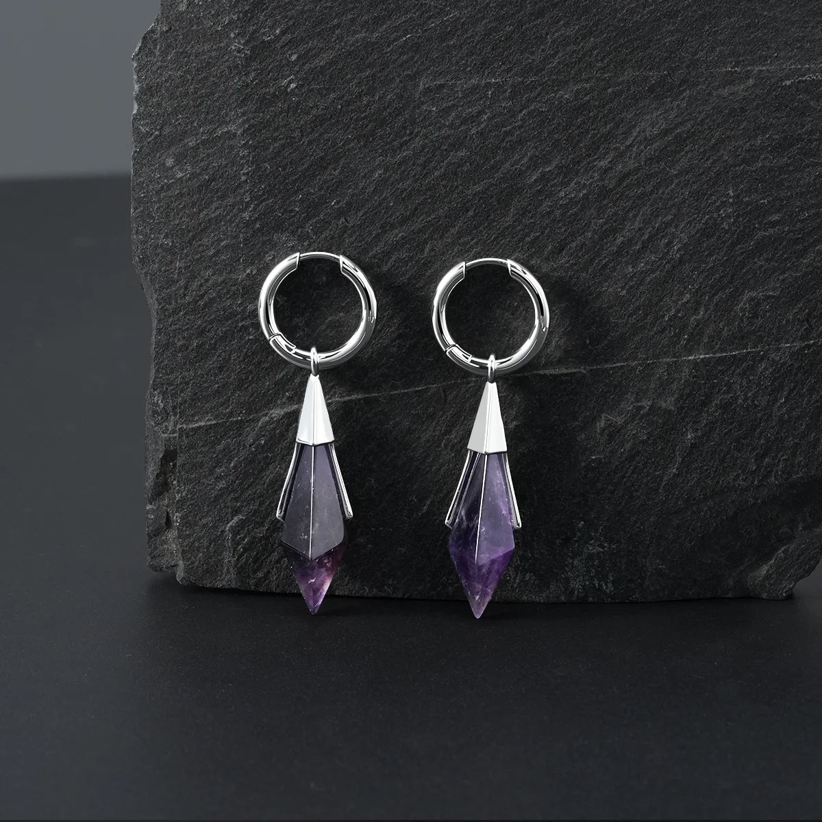 Silver Prism Apex Earrings displayed on a dark stone surface, featuring faceted amethyst crystal drops and polished hoop design.
