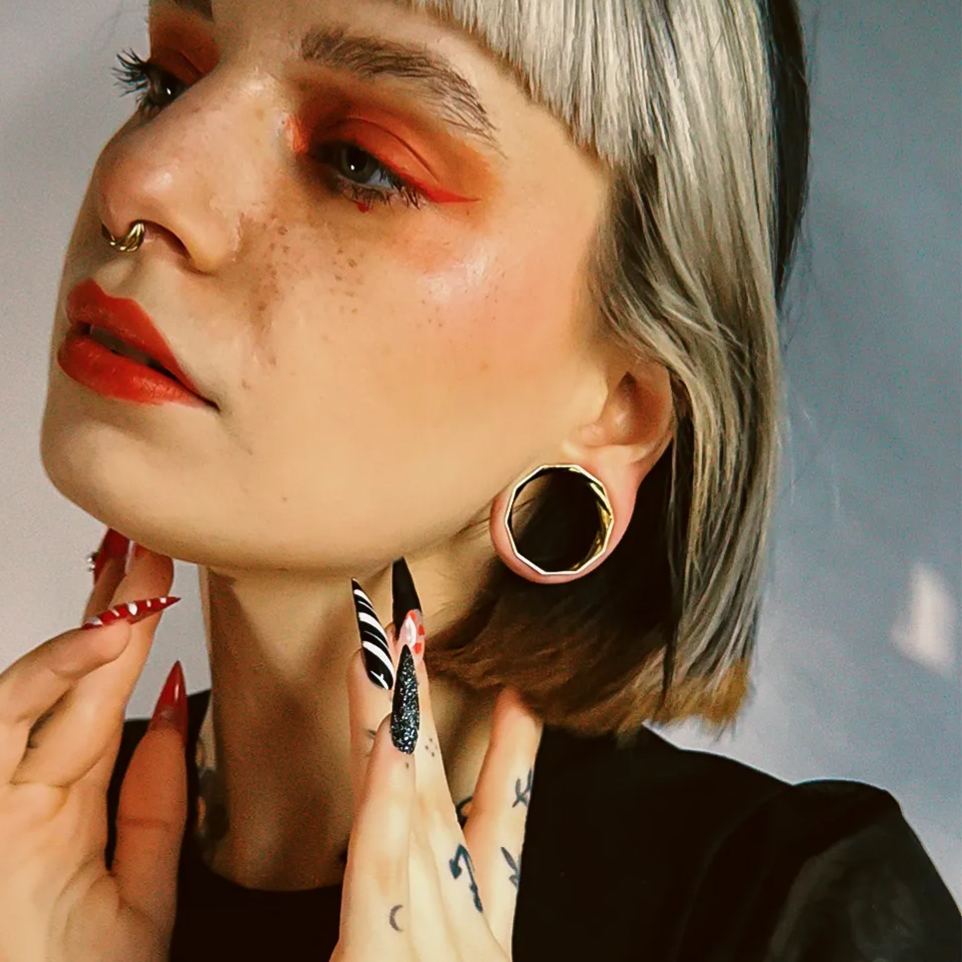 Close-up of a model wearing a faceted gold-tone tunnel earring with orange eye makeup and styled bob