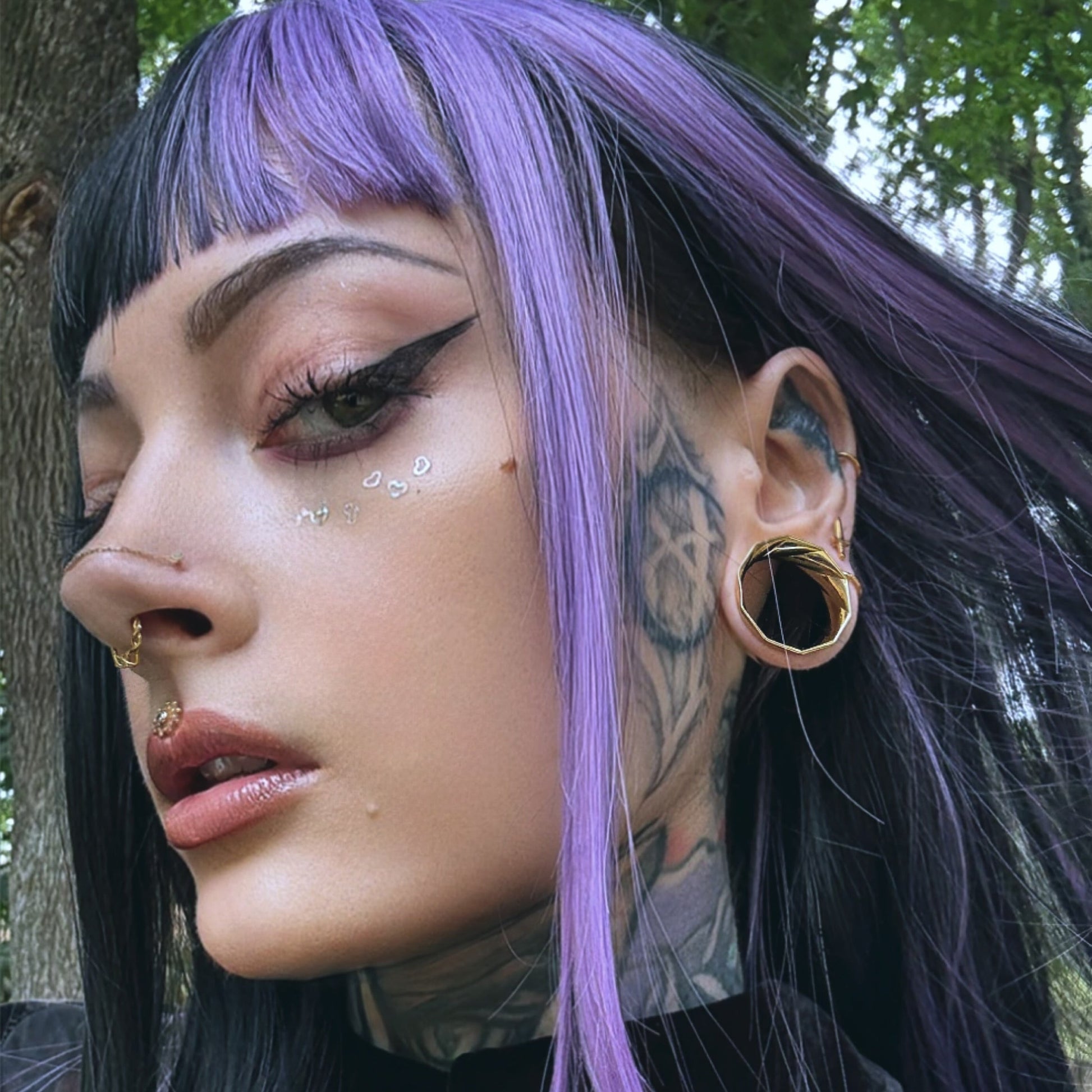 Close-up of a model with purple bangs wearing a twisted gold-tone tunnel earring and layered nose rings