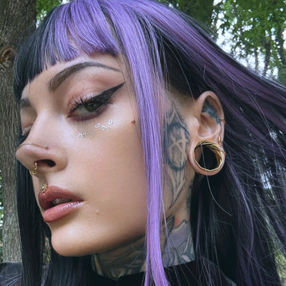 Close-up of a model with purple bangs wearing a twisted gold-tone tunnel earring and layered nose rings