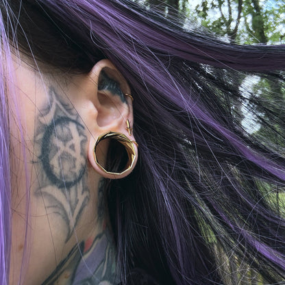 Side profile of a model with purple hair and neck tattoos wearing a faceted gold-tone tunnel earring