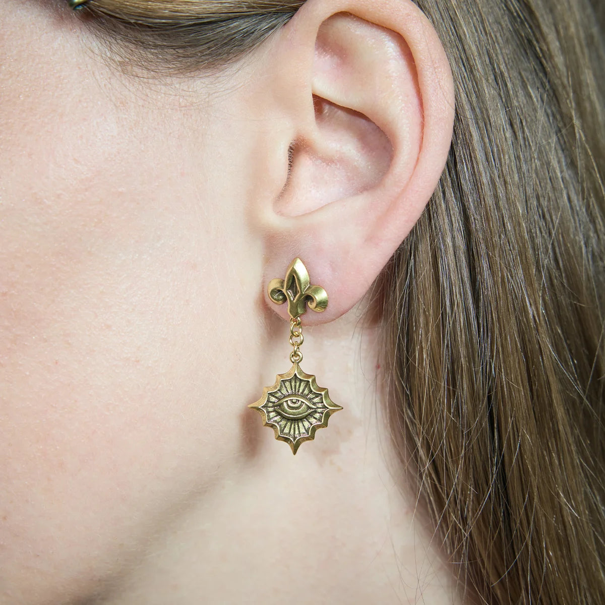 Close-up of a gold-tone Radiant Witness Drop Earring worn from the side, highlighting the fleur-de-lis stud and radiant eye medallion.
