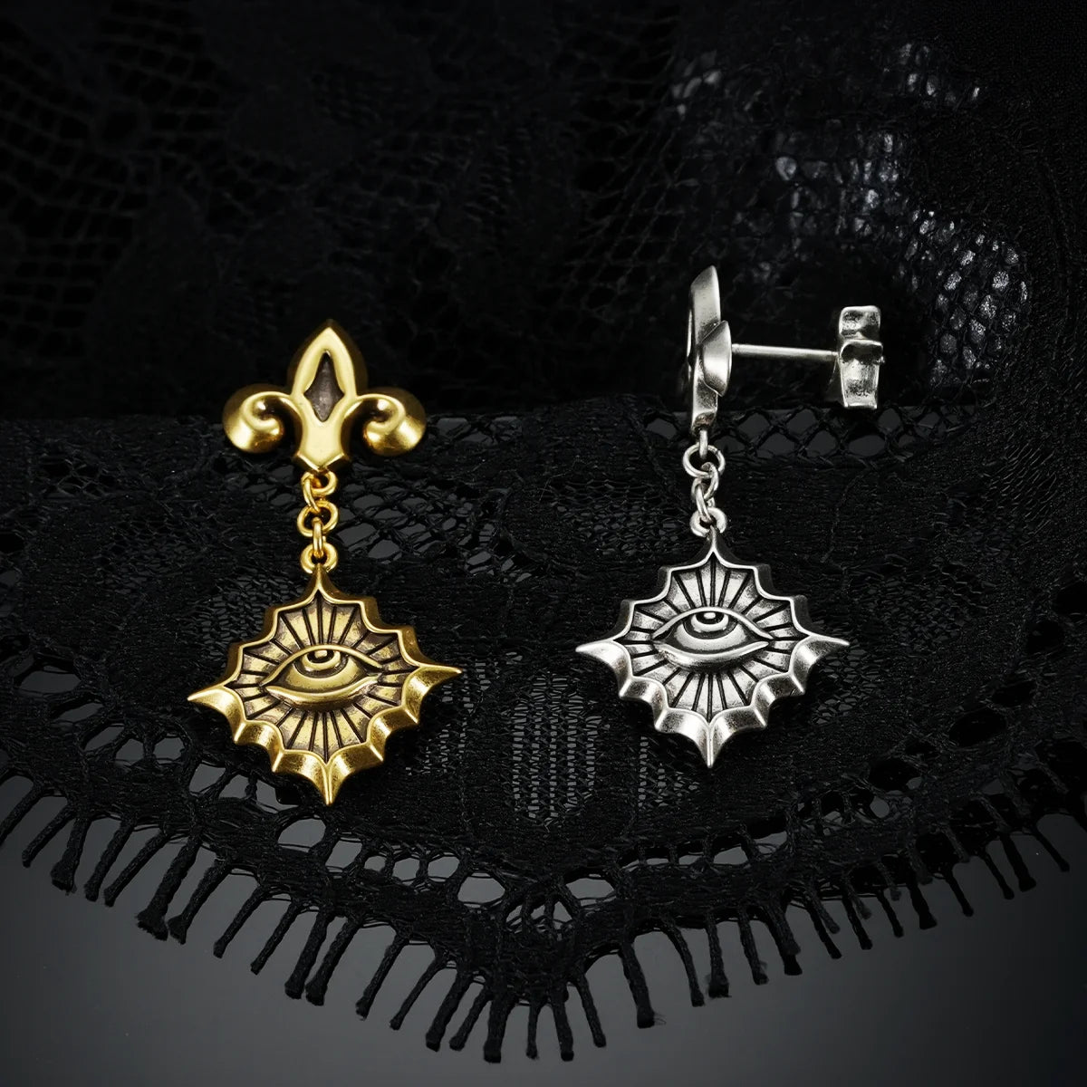 Antiqued silver- and gold-tone Radiant Witness Drop Earrings displayed together against black lace on a dark reflective surface.