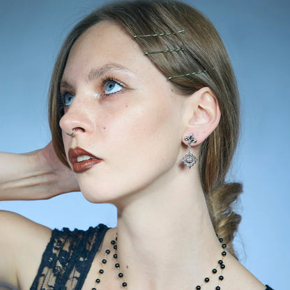 Low-angle portrait showing antiqued silver-tone Radiant Witness Drop Earrings, with one hand resting behind the neck.