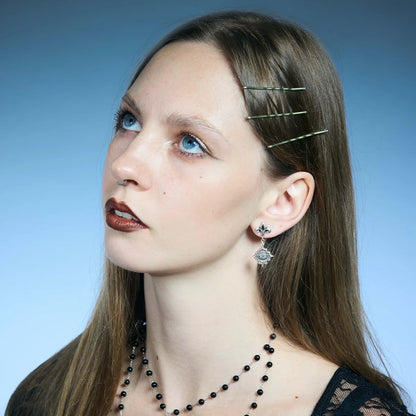 Low-angle portrait showing antiqued silver-tone Radiant Witness Drop Earrings suspended below fleur-de-lis studs.