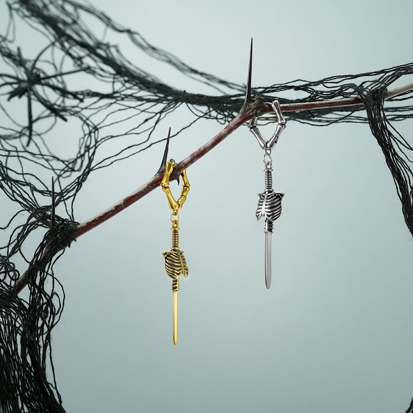 Silver and gold Ribcage Blade earrings hanging from a dark branch