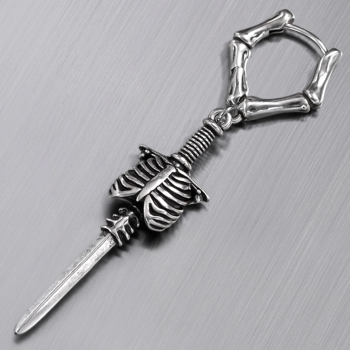 Close-up of a silver-tone bone hoop earring with ribcage and blade charm on textured surface