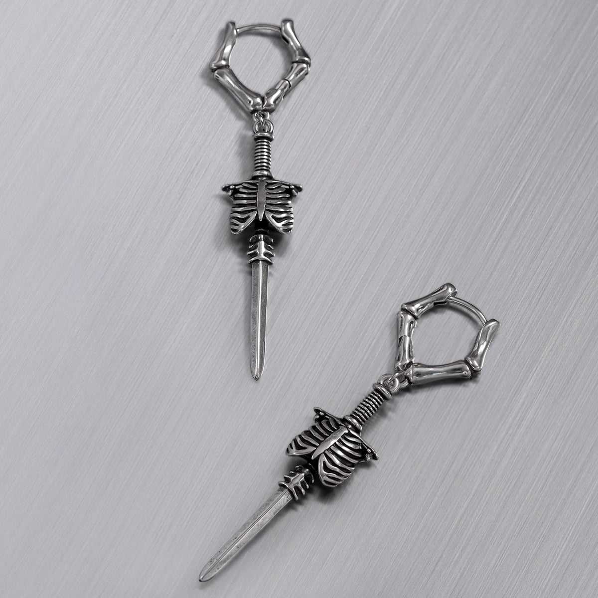 Pair of silver-tone bone hoop earrings with ribcage and blade charms arranged on textured surface