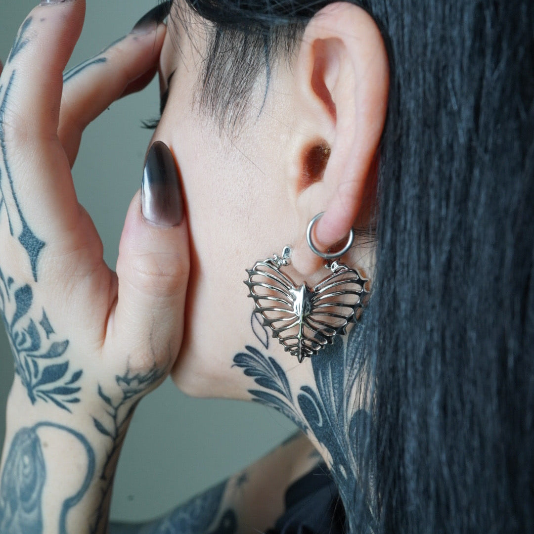 Silver-tone ribheart earring worn on tattooed model, close-up side view showing heart rib cage motif