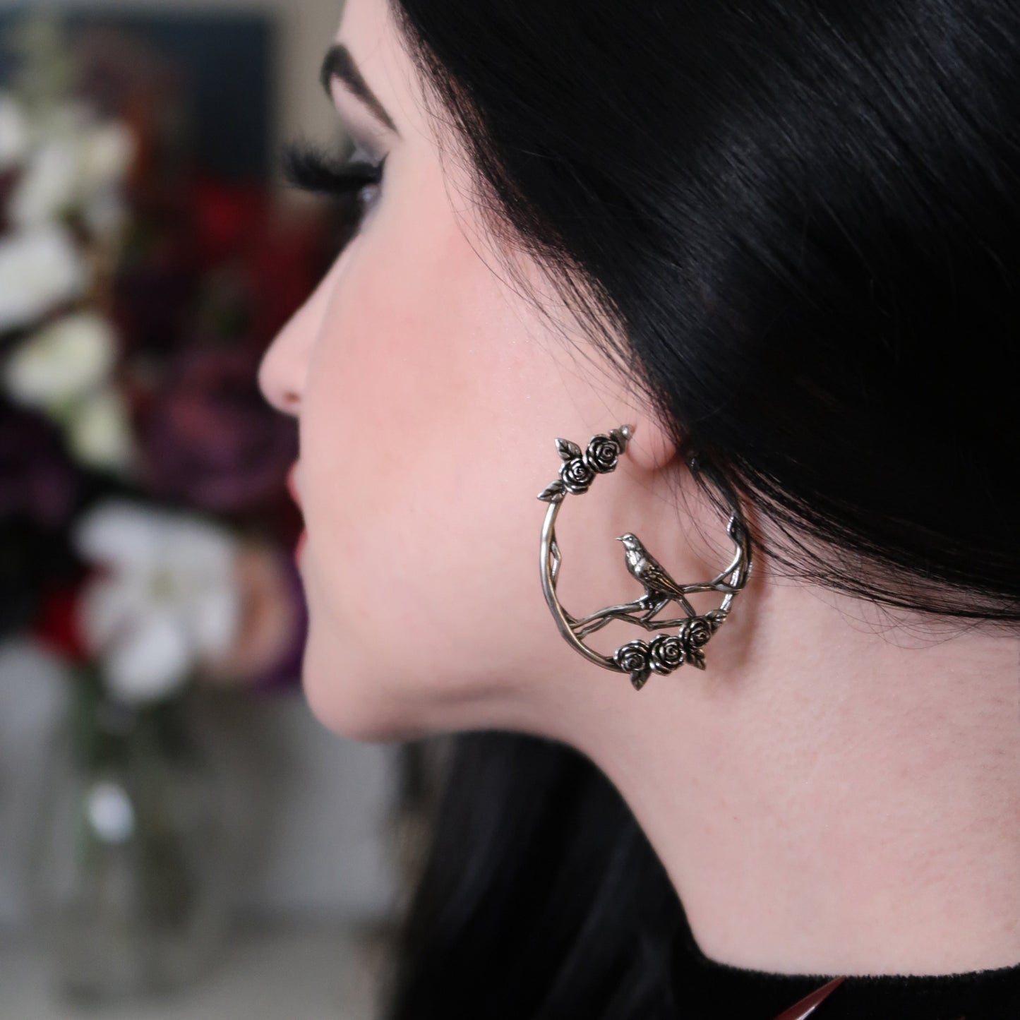 Dark-haired model wearing a silver-tone hoop earring featuring roses and a bird motif