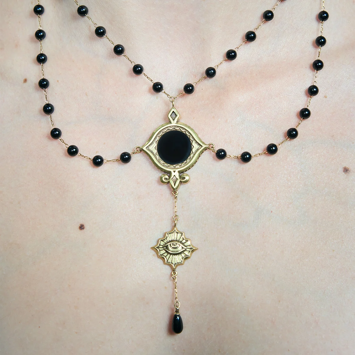Close-up of a gold-tone Scrying Mirror Necklace worn at the chest with an oval black obsidian centerpiece and descending eye charm.