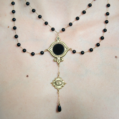Close-up of a gold-tone Scrying Mirror Necklace worn at the chest with an oval black obsidian centerpiece and descending eye charm.