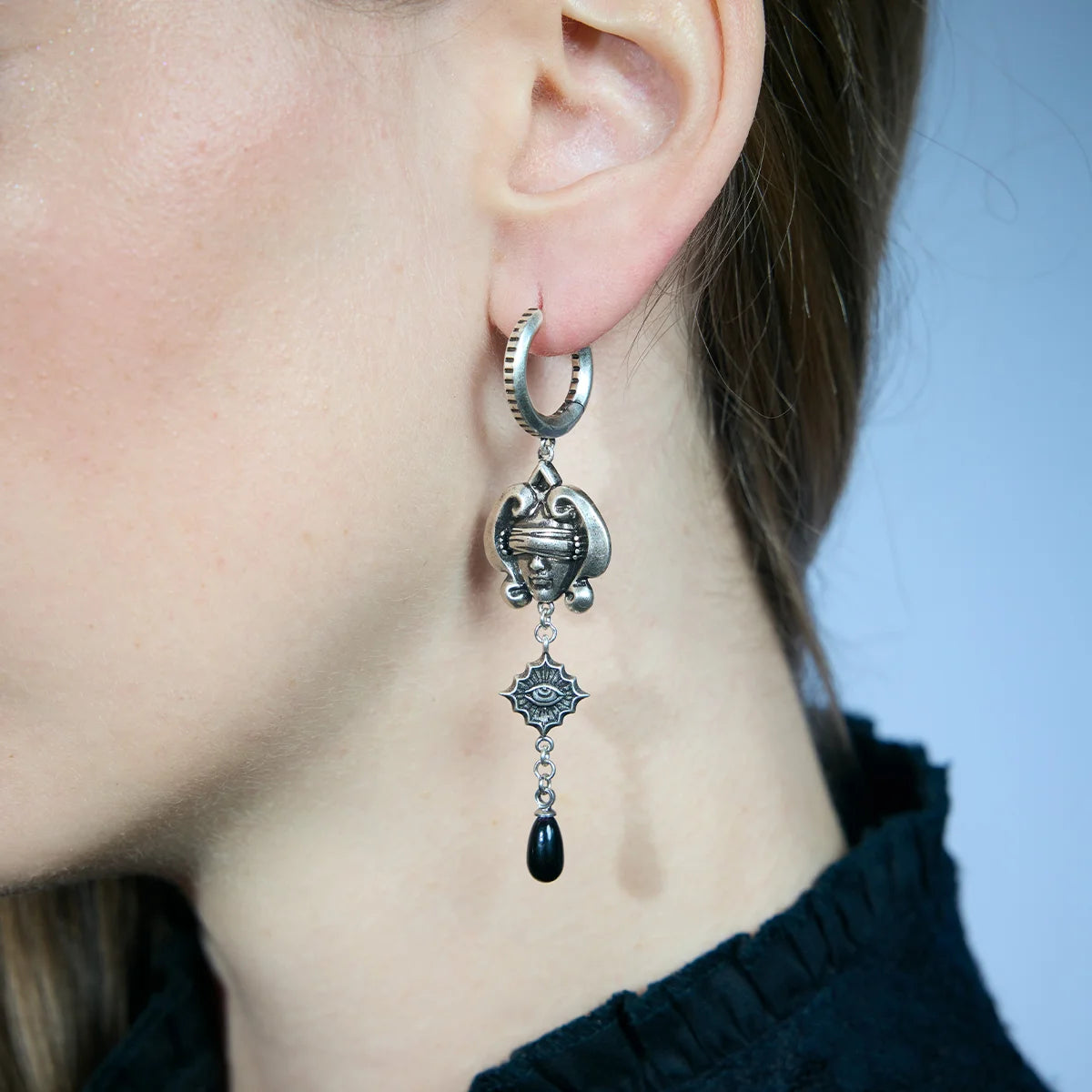 Close-up of an antiqued silver-tone Second Sight Drop Earring worn from the side, showing the blindfolded face, eye medallion, and black teardrop.