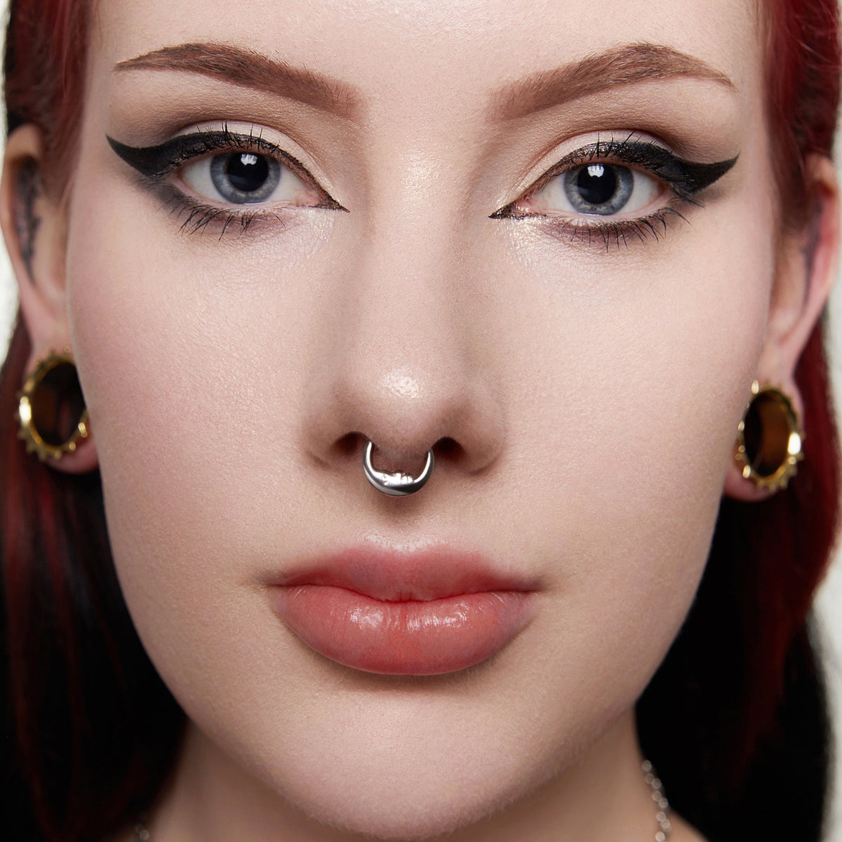Close-up of a model's face wearing a titanium septum ring alongside matching gold ear plugs