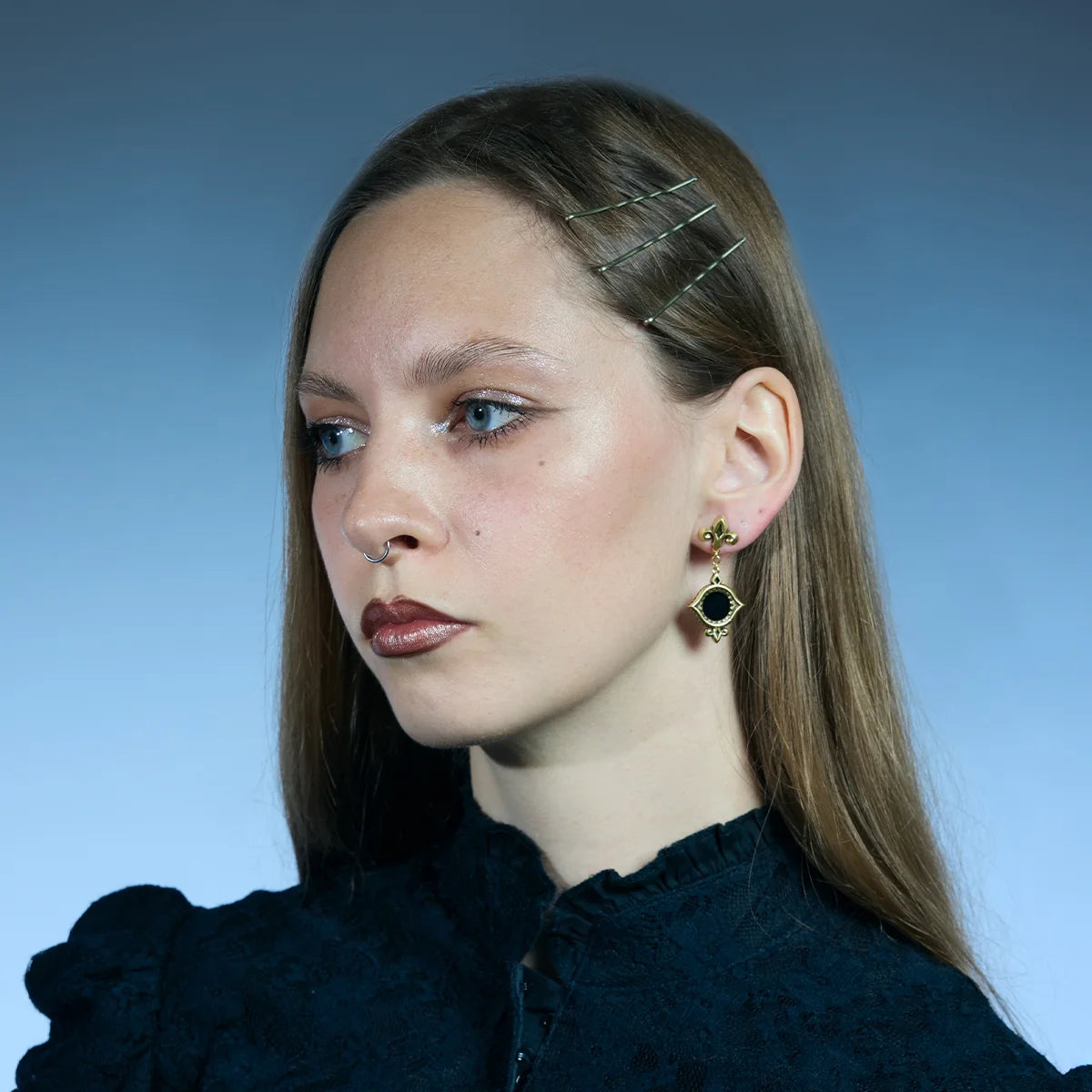 Three-quarter portrait showing gold-tone Silent Lens Drop Earrings with round black obsidian centers and fleur-de-lis studs.