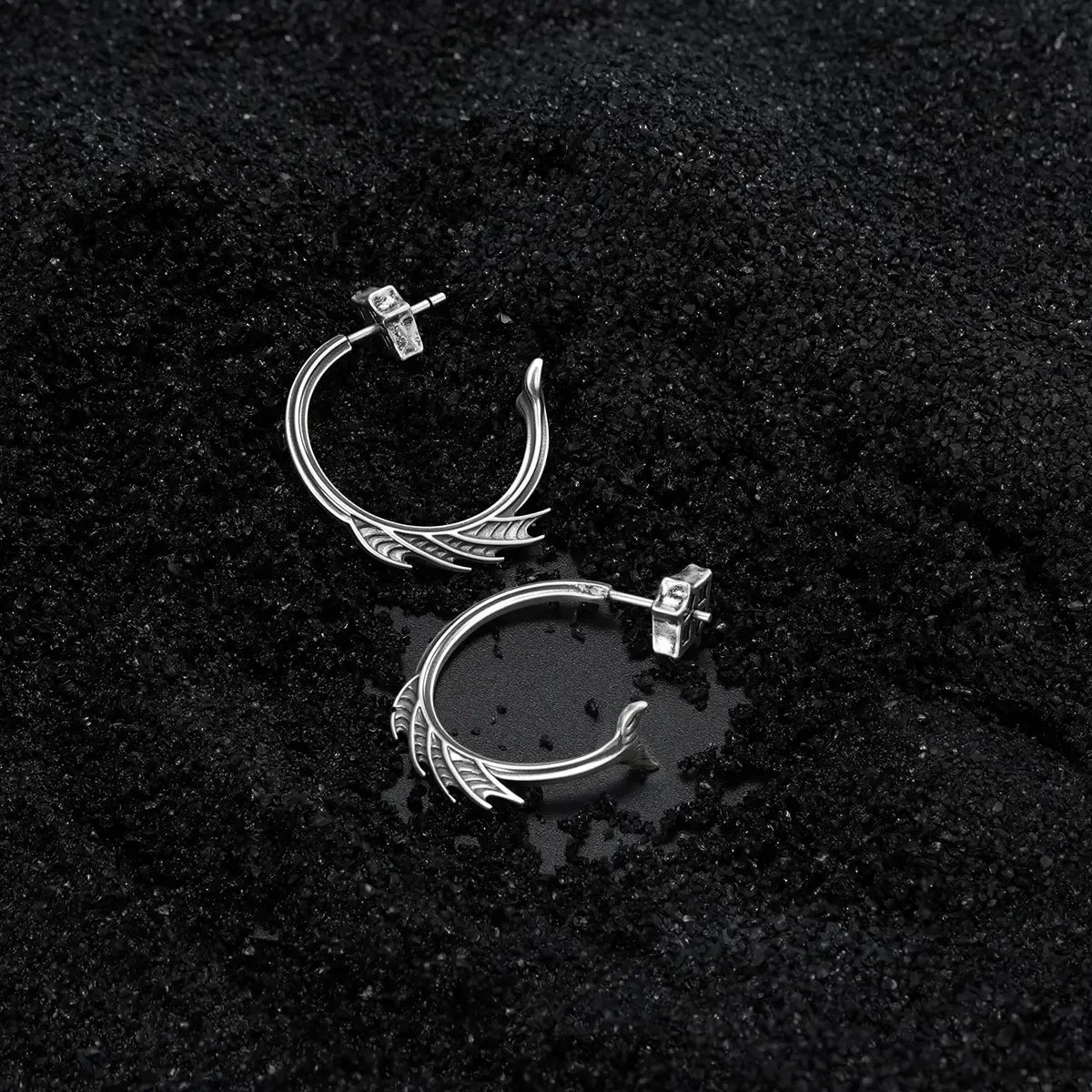 Silver Siren Crest Earrings displayed on black sand, highlighting the sculptural half-hoop shape and ocean relic mood.
