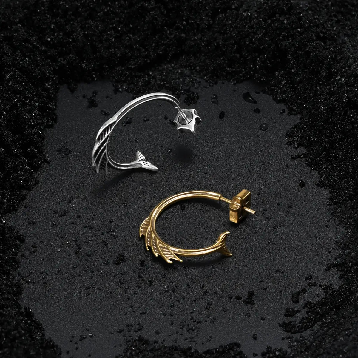 Silver and gold Siren Crest Earrings displayed on black sand, featuring curved half-hoop shapes and sharp fin-like ridge details.
