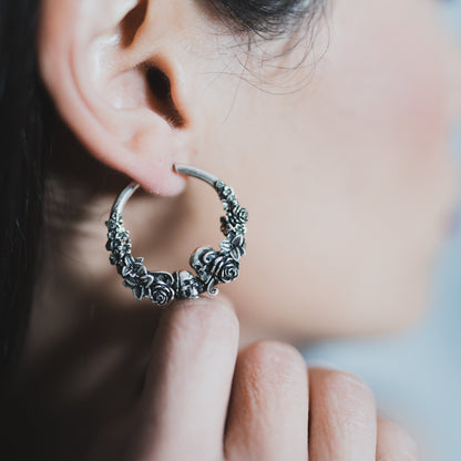 Close-up of an oxidized silver-tone hoop earring with skull and rose detailing worn on the ear, fingers touching it