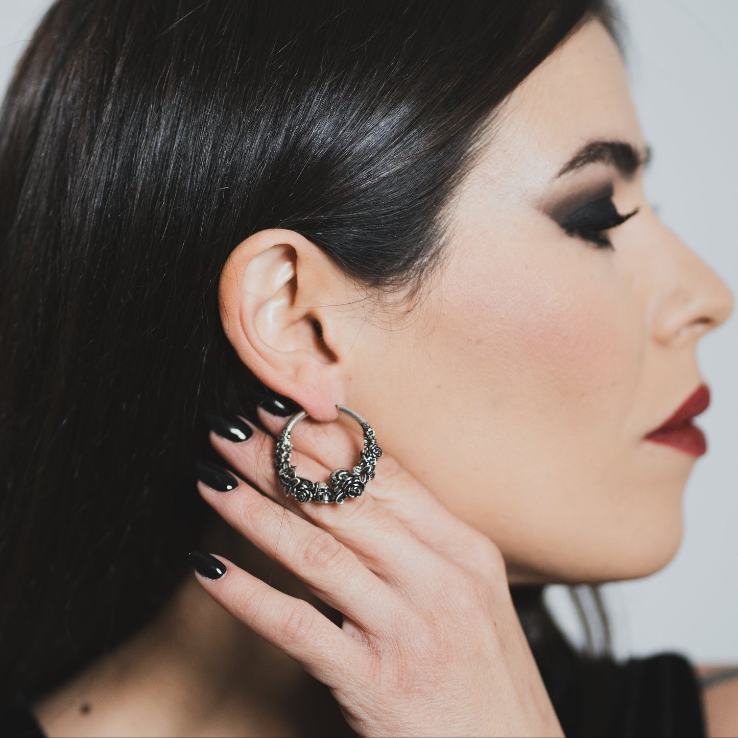 Model with dark makeup wearing an oxidized silver-tone skull and rose hoop earring, close-up side profile view
