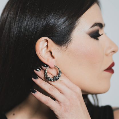 Model with dark makeup wearing an oxidized silver-tone skull and rose hoop earring, close-up side profile view