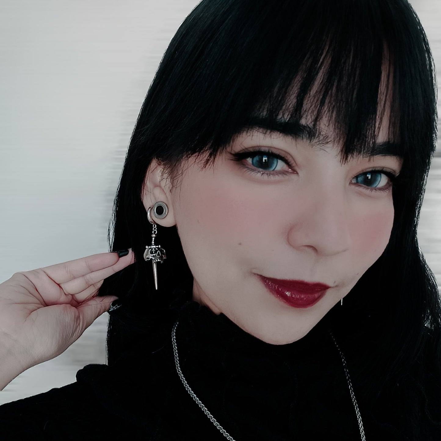Model with dark hair wearing a silver-tone skull dagger drop earring, close-up portrait