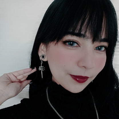 Model with dark hair wearing a silver-tone skull dagger drop earring, close-up portrait