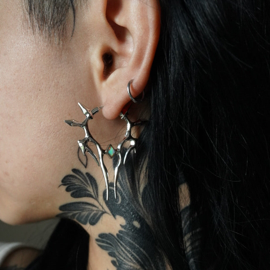 Silver-tone spiked hoop earring with a small green stone, worn close-up on a model with dark hair and neck tattoo