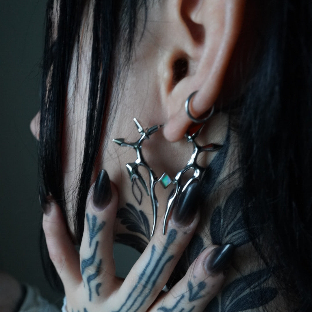 Close-up of a silver-tone spiked earring worn on the ear as a model with tattooed hands touches the face