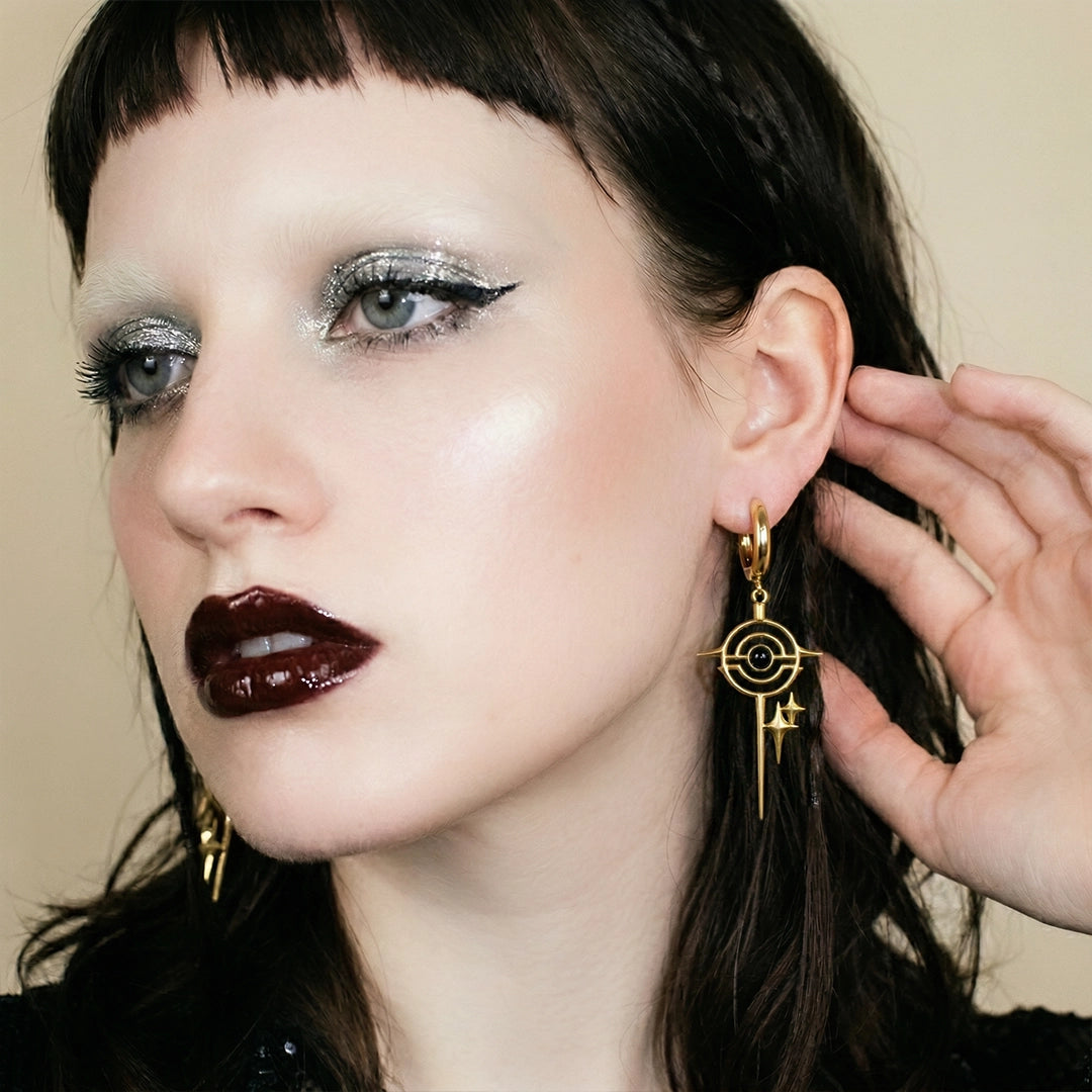 Model wearing gold-tone drop earrings with a circular star motif and black stone, hand near ear