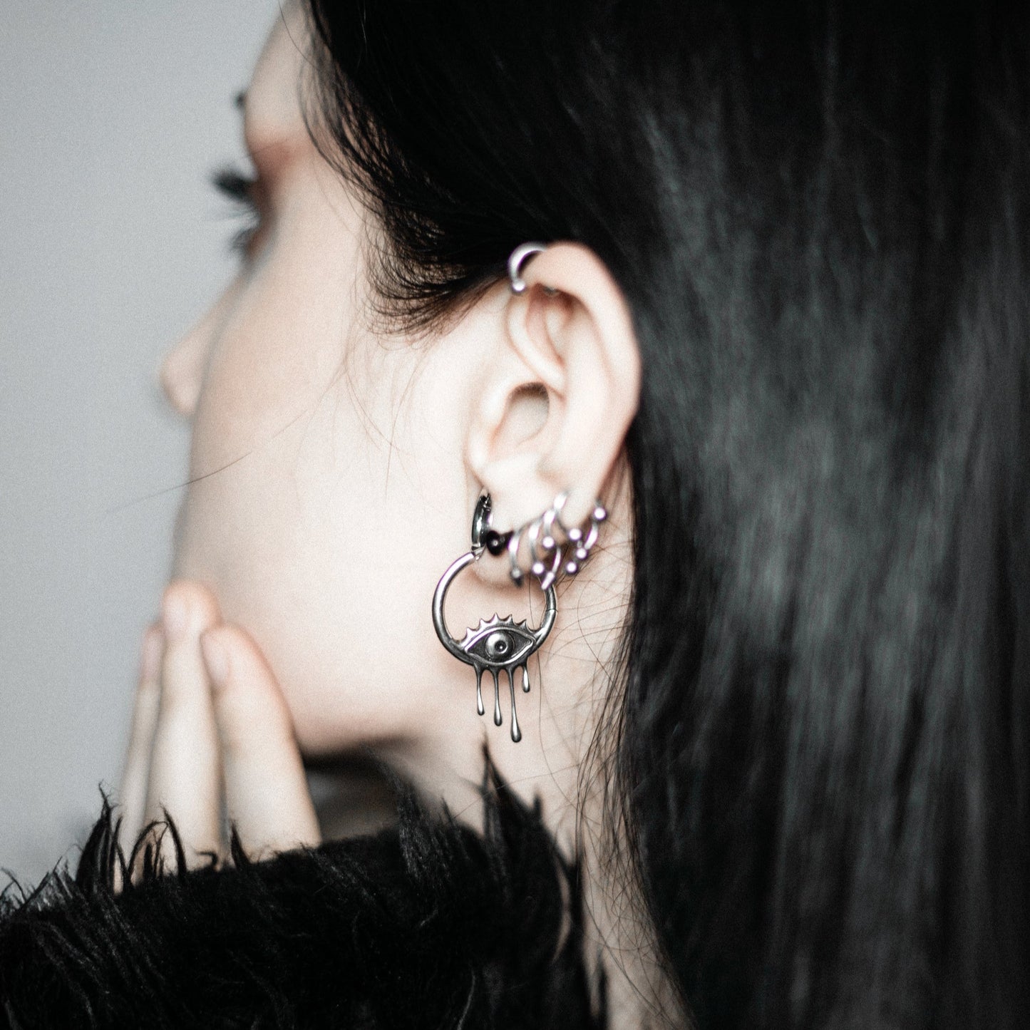 Side view of a model with dark hair wearing a silver-tone hoop earring with a dripping eye charm and small hoop studs