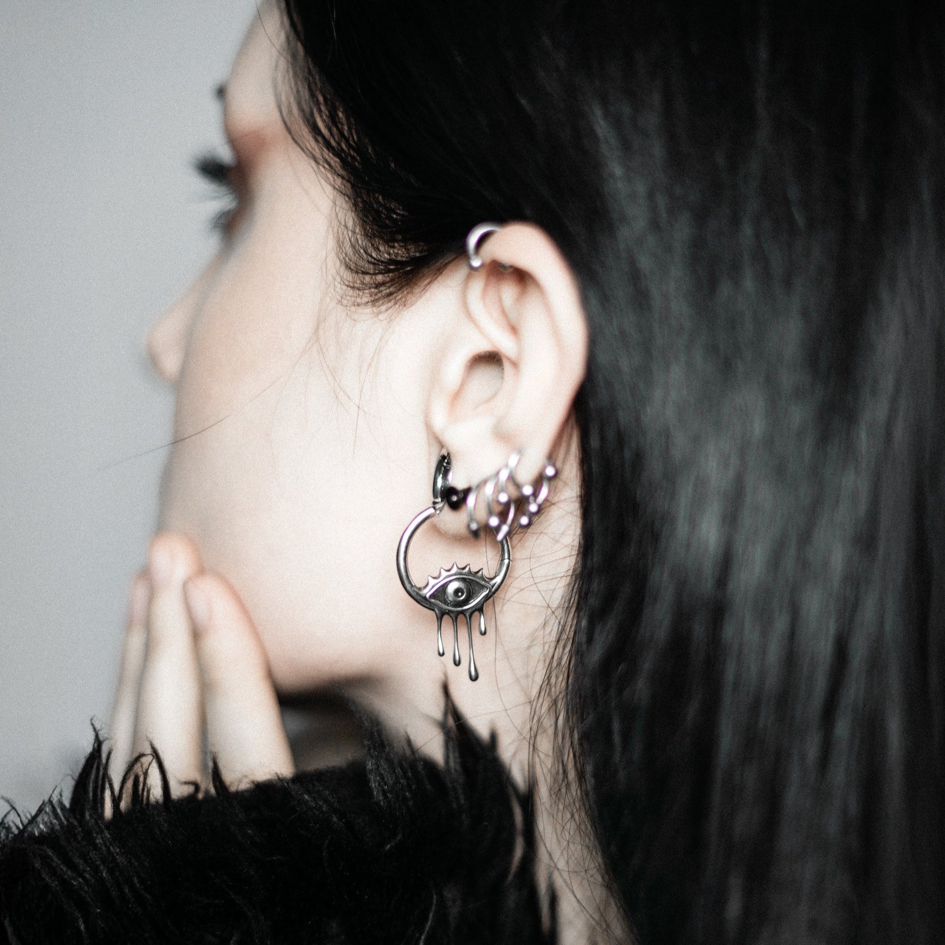 Side view of a model with dark hair wearing a silver-tone hoop earring with a dripping eye charm and small hoop studs