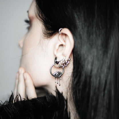 Side view of a model with dark hair wearing a silver-tone hoop earring with a dripping eye charm and small hoop studs