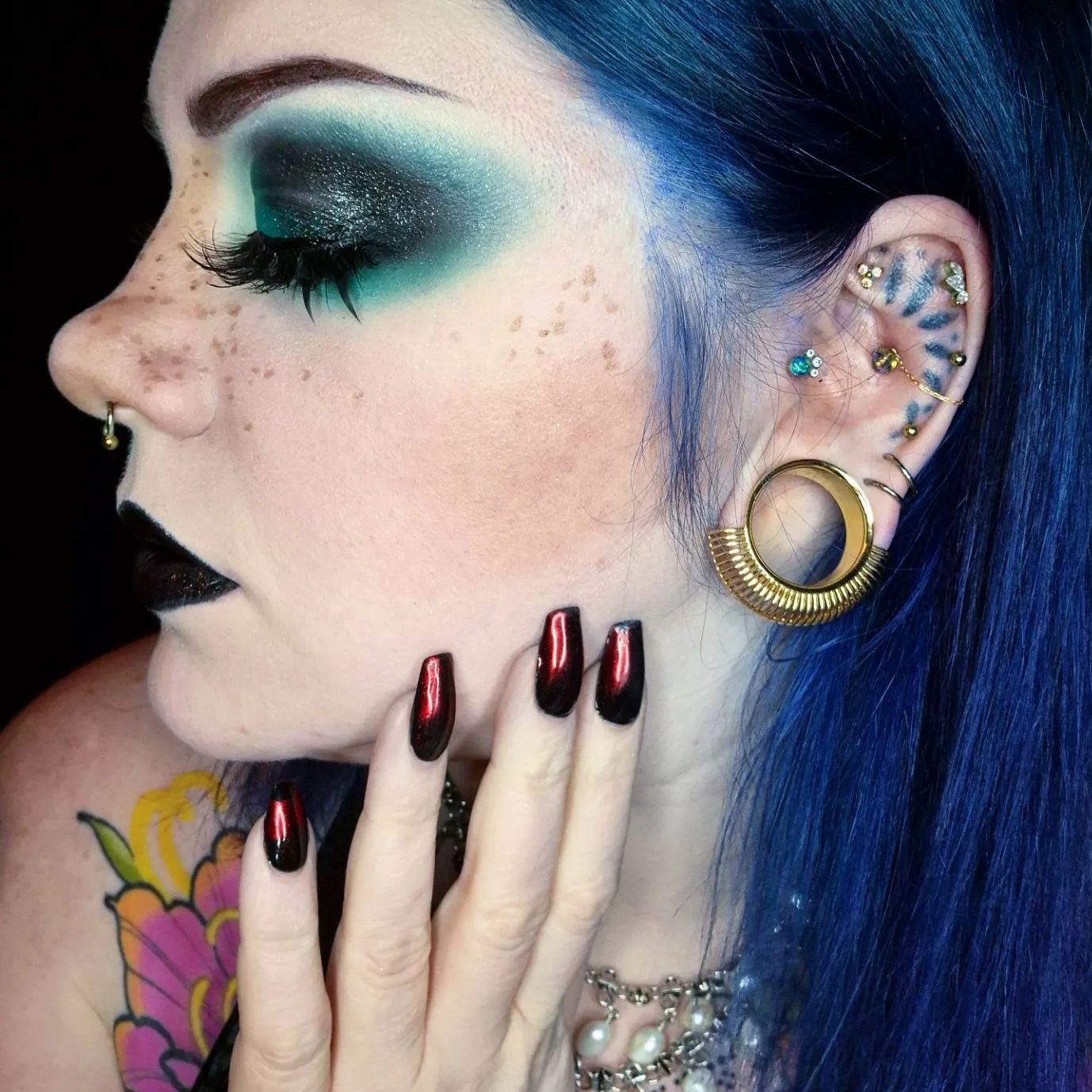 Model with blue hair and dark makeup wearing a gold-tone sunburst tunnel with several ear piercings