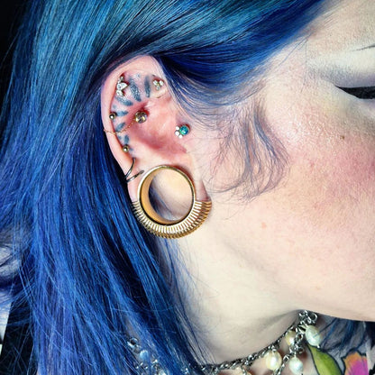 Ear with blue hair showing a gold-tone radiance tunnel worn alongside multiple small stud piercings
