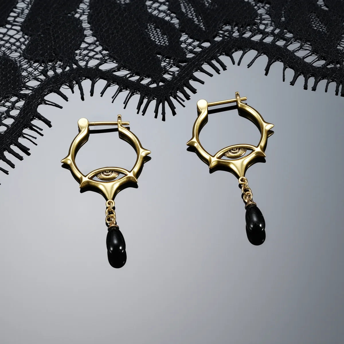 Antiqued silver- and gold-tone Thorned Gaze Hoop Earrings displayed together against black lace on a dark reflective surface.