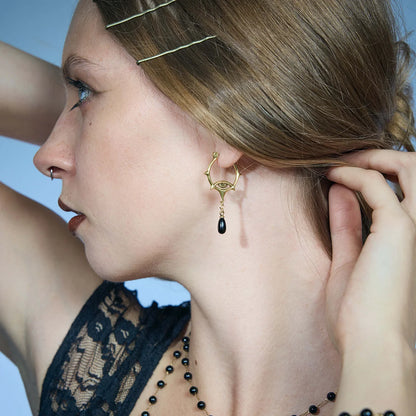 Close-up of a gold-tone Thorned Gaze Hoop Earring worn from the side with outward-facing thorns and a suspended black teardrop accent.